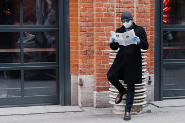 Full length shot of serious man dressed in black coat, wears sunglasses and medical mask, reads...