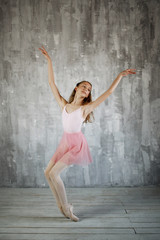 Fototapeta na wymiar Beautiful female flexiable ballet dancer doing exercise on a gray background. Adorable ballerina is wearing a pink leotard and skirt, beige stockings, pointe shoes.
