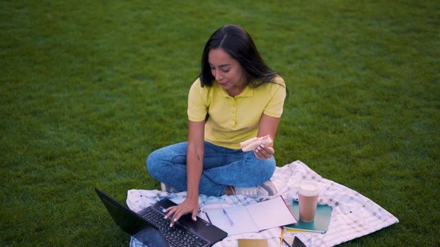 Campus, concentrated girl student doing homework. Girl works on laptop and eats a sandwich. 4K