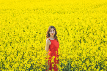 Naklejka na ściany i meble Young beautiful girl in a red dress close up in the middle of the yellow field with the radish flowers closeup