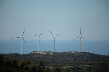 White wind turbines that generate electricity from renewable sources of nature, surrounded by hills and trees on a sunny day. Eco green energy.