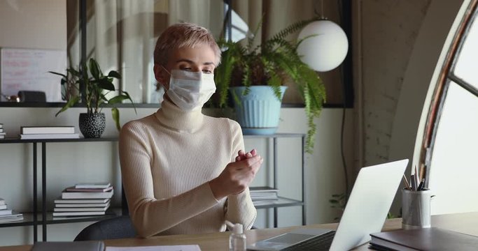 Female worker wears mask cleaning hands with sanitizer gel working from home office sits at desk. Businesswoman employee using alcohol sanitiser before using computer to prevent coronavirus concept.
