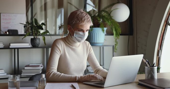 Young business woman wearing protective face mask and medical gloves working on laptop computer sitting at home office desk. Female worker doing distance remote job on corona virus quarantine concept.