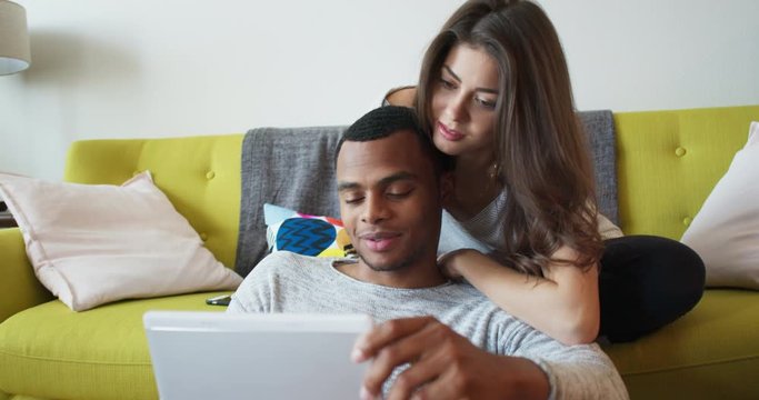 Young millennial couple sitting on couch in living room using tablet computer together. African American and Caucasian boyfriend and girlfriend using device online. 4k Slow motion handheld