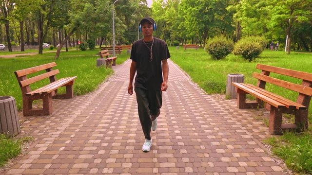mixed race young man with headphones listening music in casual trendy clothes and sneakers walking to the camera on the street slow motion summer landsca