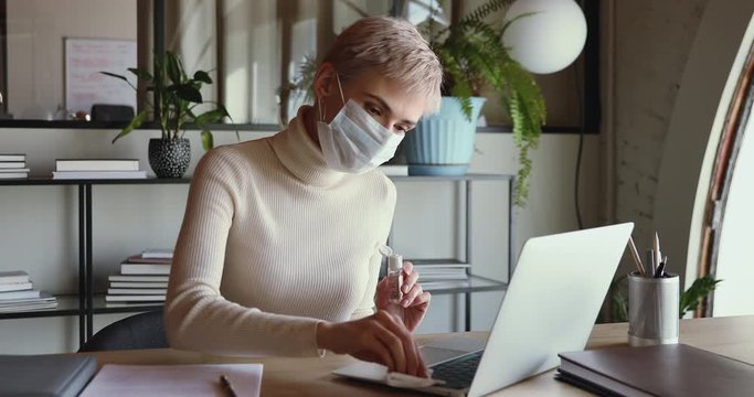 Young business woman wears medical face mask cleaning laptop keyboard with sanitizer to stop coronavirus spreading. Female worker using sanitiser gel disinfecting workplace working from home office.
