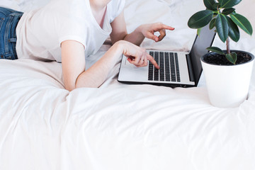 home office in bed. young woman wearing white shirt and blue jeans working at home on white bed sheets with a white laptop and potted plant