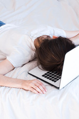 home office in bed. young woman wearing white shirt and blue jeans and sleeping on a white laptop in bed with white sheets