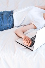 home office in bed. young woman wearing white shirt and blue jeans and sleeping on a white laptop in bed with white sheets