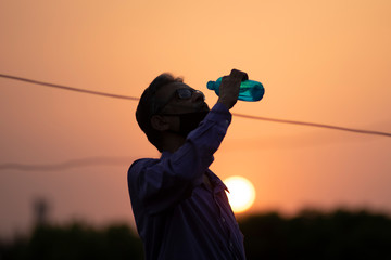 Silhouette portrait of an Indian old man with corona preventive mask drinking water on a rooftop during sunset in home isolation.Indian lifestyle, disease and home quarantine.