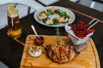 grilled pork chop with raspberry sauce and grilled garlic, potato, pumpkin vegetable on wooden board served with potato chips in basket. with salmon salad and orange mix cold brew coffee on table.
