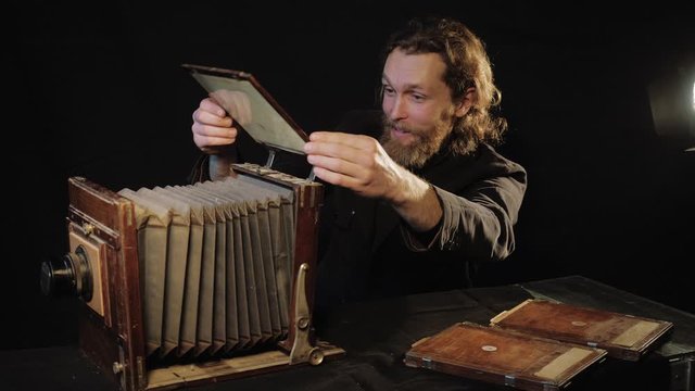 Enthusiastic and cheerful bearded man, photographer in image of Dostoevsky, examines and inserts photographic plates into old 19th-century antique wooden large plate camera in room black background