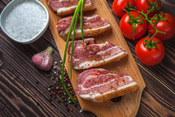 Raw slices of pork bacon on a cutting board, on a rural wooden tabletop. Pork belly with vegetables.
