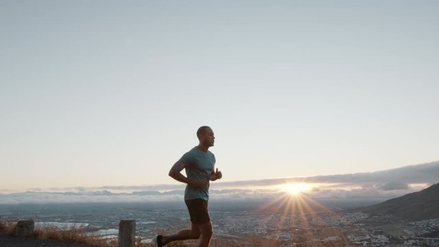 Young man running with the sun behind him on a mountain road. Man on a morning run on a hill road with the city in background.
