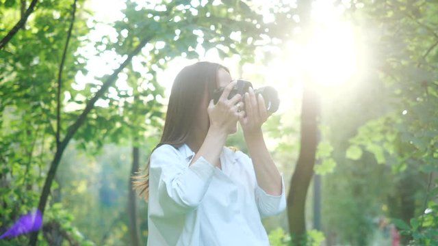 Young beautiful girl with long hair takes pictures on a professional camera. Attractive woman in a white shirt photographs the nature of a cozy city park on a warm sunny day in spring, middle view.