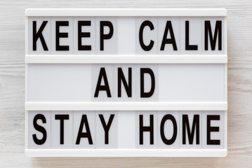 Fototapeta premium 'Keep Calm and Stay Home' words on a lightbox on a white wooden background, top view. Overhead, from above, flat lay.