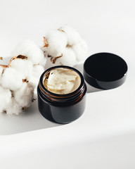 A black glass jar with a cosmetic cream for face or body on white background with cotton. Selective focus