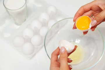 Close-up on the hands of a woman who cracks an egg with her fingers and pours the yolk into a plate against the background of a package with a dozen eggs and a glass with flour