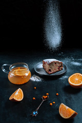 Tasty cake with flying icing sugar and a teapot of sea buckthorn tea with oranges on a dark background.