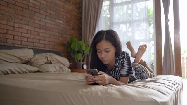 happy smiling teenage asian girl using smartphone at home while laying on the bed