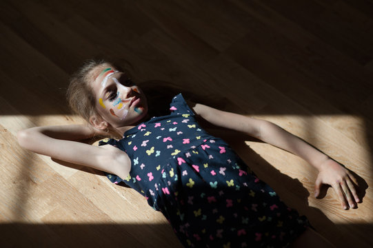 Beautiful Little Girl With Face Art Painted With Colors In Blue Dress Lying On Floor At Home During Social Distancing Self Isolation