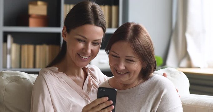 Head shot smiling young woman showing funny mobile application to middle aged mum, resting together on sofa. Happy mature mother posing for selfie shot on cellphone with grown up daughter at home.