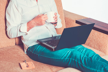 Naklejka premium young man in white shirt and blue jeans hold cup of tea and working on laptop, writes in notebook in home office sitting on cozy beige sofa Stay at home, online work, distance learning concept