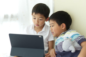 Two little young Asian boys are playing and watching the digital tablet at home for the game,e-learning and education with fun and joy during day time next to the window curtain.