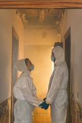 Couple of virologist doctors and scientists wearing biohazard protective suits and holding hands in a contaminated house by coronavirus outbreak