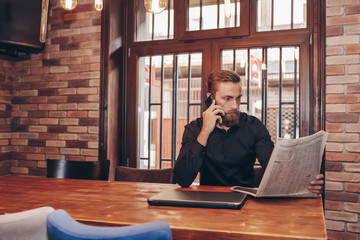 Bearded young businessman reading a newspaper and talking on a mobile phone