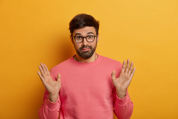 Do not involve me. Puzzled bearded man raises palms as dont want to got into trouble, bother with someone else, looks hesitant and confused, wears optical glasses and pink jumper, poses indoor