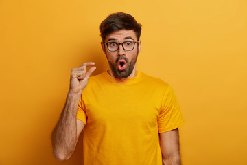 Deeply impressed bearded man shocked by very tiny size, measures something small, gasps from amazement, has unexpected reaction on decreased price, stares through glasses, wears yellow t shirt - obrazy, fototapety, plakaty