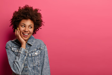 Portrait of interested cheerful Afro American woman looks aside with broad smile, feels happy, touches cheek, dressed in stylish denim jacket, isolated on pink background. Copy space for promotion