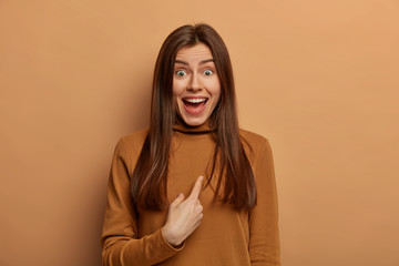 Excited positive European woman points at herself with happiness and surprise, being told good news, asks question, dressed in brown turtleneck, isolated on beige wall, shocked to hear her name