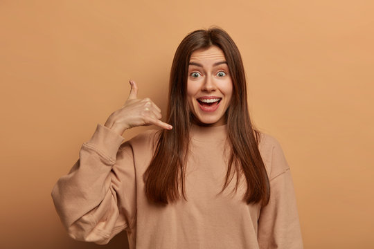 Communcation on distance concept. Happy European woman makes phone gesture, asks telephone number or give her call, smiles joyfully, dressed in casual sweatshirt, isolated on brown background.