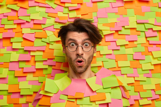 Emotional young European man gasps from fear, stands speechless, hears exciting incredible news, cannot believe, wears round spectacles, poses through paper background with colorful sticky notes