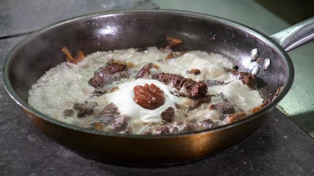 Chef in restaurant kitchen mixes ingredients in frying pan with wooden spoon mushrooms, onions, wild deer meat with cream and red sauce. Cooking for visitors. dish is fried and boils on stove.