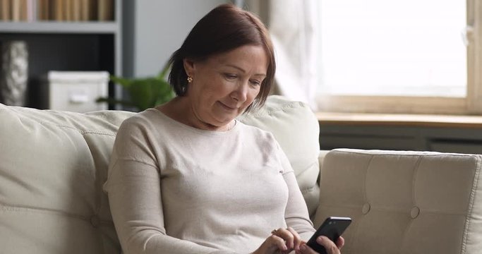 Happy middle aged brunette woman sitting on sofa, using mobile applications. Smiling elderly mature retired lady chatting in social networks with friends, watching funny photos videos easy tech usage.
