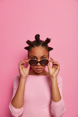 Fashion model with bun hairstyle looks seriously through sunglasses, wears rosy jumper, stands alone, going to have walk during sunny day, poses against pink background, copy space above for text