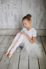 A little adorable young ballerina in white dress trying on her point shoes sitting on woody floor and gray studio background
