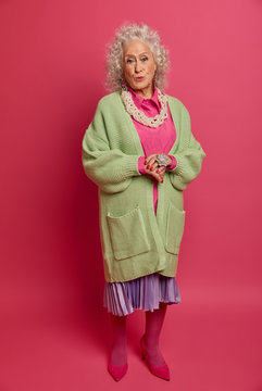 Full Length Shot Of Fashionable Mature Lady Dressed In Stylish Jumper, Pleated Skirt And Shoes, Pouts Lips And Looks With Romantic Expression At Camera, Isolated On Pink Wall. Retired Woman Indoor