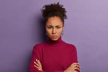 Fototapeta na wymiar Headshot of displeased dark skinned woman keeps arms folded, has offended look, frowns face, expresses negative attitude, has curly combed hair, wears burgundy poloneck jumper, poses indoor.