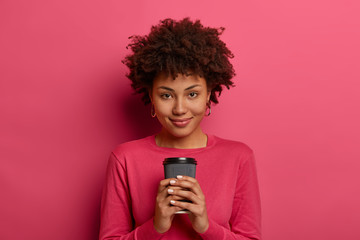 Young pretty woman with Afro haircut, holds disposable paper cup of coffee, looks directly at camera with satisfied expression, enjoys free time for break, dressed casually. Female with hot beverage