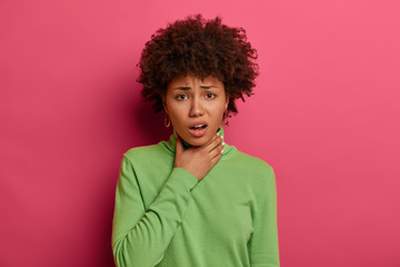 Fototapeta na wymiar Sick dark skinned young woman feels unwell, suffers from sore throat, has suffocation and cough, frowns face, wears green turtleneck, feels pain after catching cold, isolated on pink background.