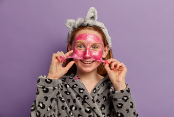 Positive small ginger European preteen girl removes face mask, enjoys perfect smooth healthy skin, wears domestic clothes, isolated on purple background. Children, beauty and wellness concept