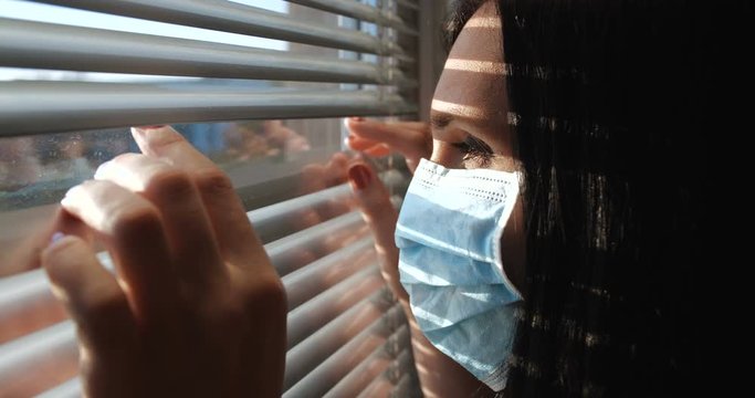 Close up front portrait from outdoors of woman wearing medical face shield pushing blinds at window with hands. Looking fearfully outside being afraid of germs. Concept of quarantine, self-isolation