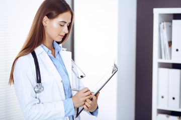 Doctor woman filling up medical form while standing near window in clinic. Physician at work. Medicine and health care concept
