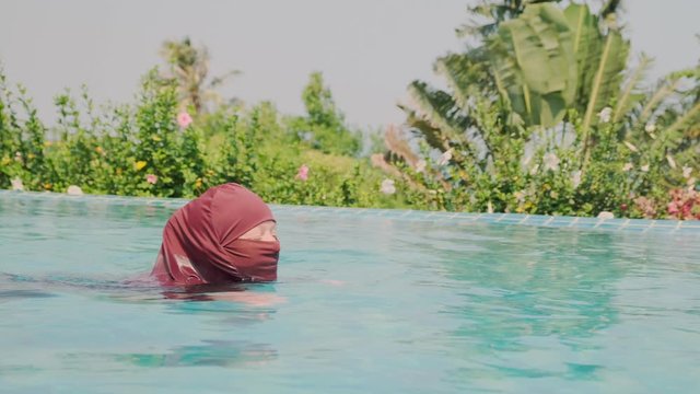 Muslim woman swims in blue water of swimming pool.