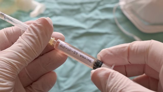 Blurred image of the sanitizing gel, a mask and a syringe. Gloved hands raise the syringe in a close-up image. It reads coronavirus vaccine, they remove the cap and let out a few drops.