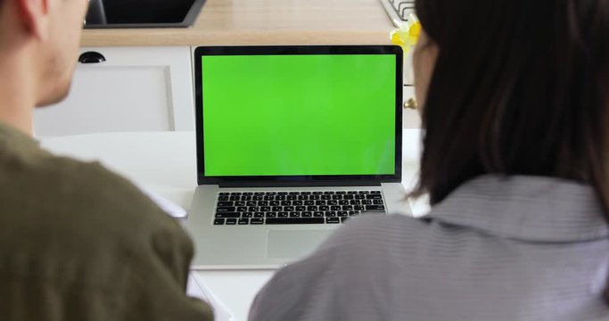 Back view of creative millennial couple working together or watching video on laptop in kitchen. European business man and woman working from home, MacBook Pro with green screen. Quarantine, COVID-19